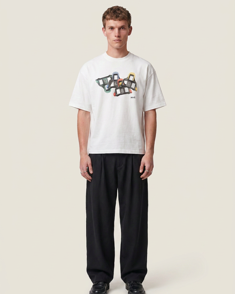 A man modeling a white short-sleeve t-shirt with a colorful geometric graphic on the chest, paired with loose black trousers and black shoes, against a solid light beige background.