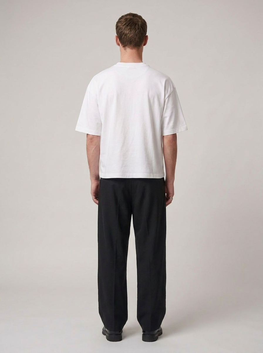 A man viewed from the back wearing a plain white short-sleeve t-shirt and loose black trousers, standing against a light gray studio background.
